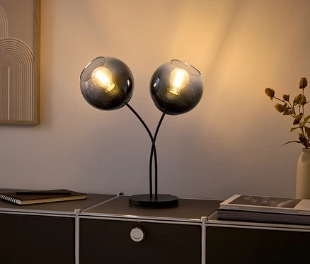 Dvě skleněné lampy s žárovkami na černé komodě.
