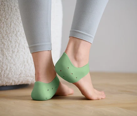 Nohy ženy v zelených silikonových návlecích na paty, stojící na dřevěné podlaze.