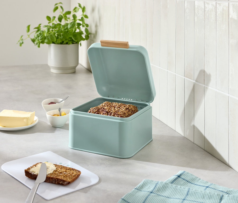 Otevřená světle modrá chlebovka s chlebem, vedle máslo, džem, nůž a namazaný chleba.