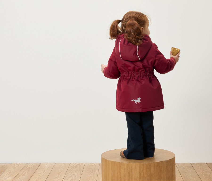 Dívka s rusými vlasy v culících stojí na dřevěném podstavci zády k fotoaparátu, má na sobě vínovou bundu s kapucí a obrázkem koně a drží v ruce kornout.