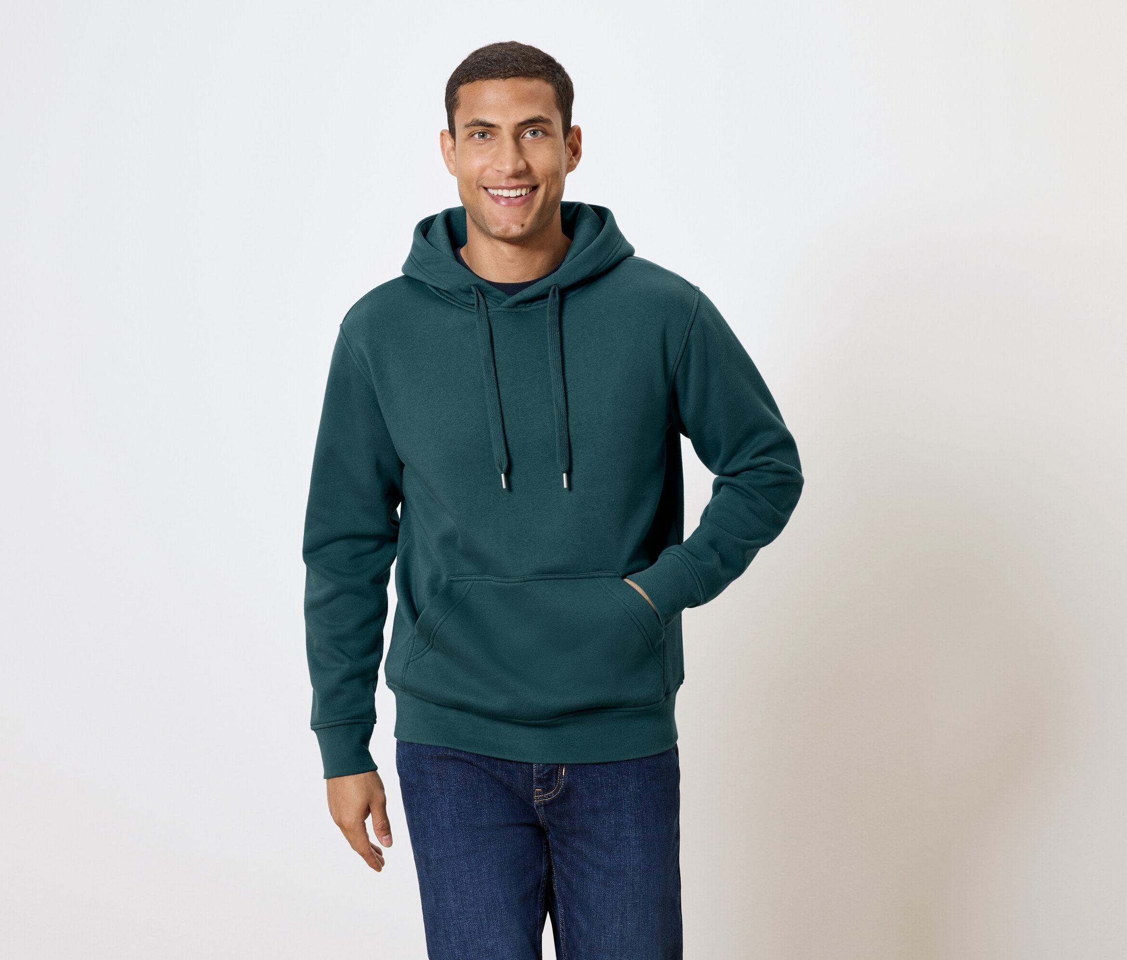 Muž s krátkými vlasy se usmívá a pózuje v tmavě zelené mikině s kapucí a v modrých džínách.