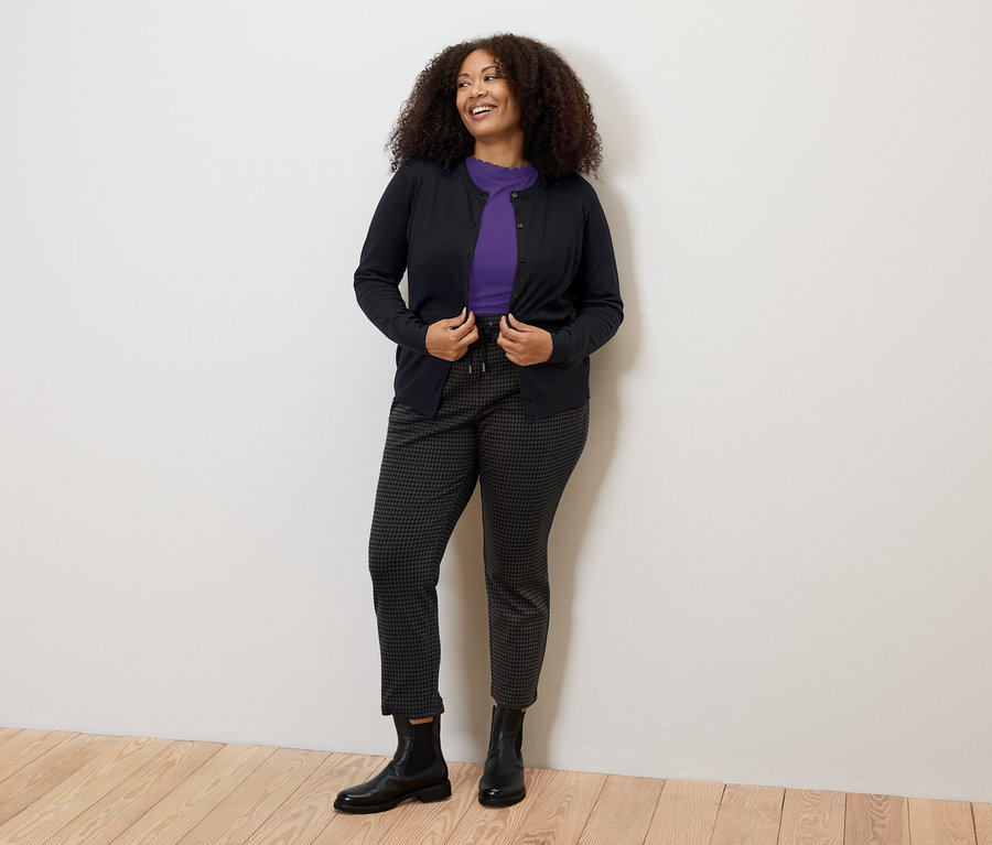 Žena má na sobě fialové tričko se zvlněným okrajem, černé kalhoty a svetr.