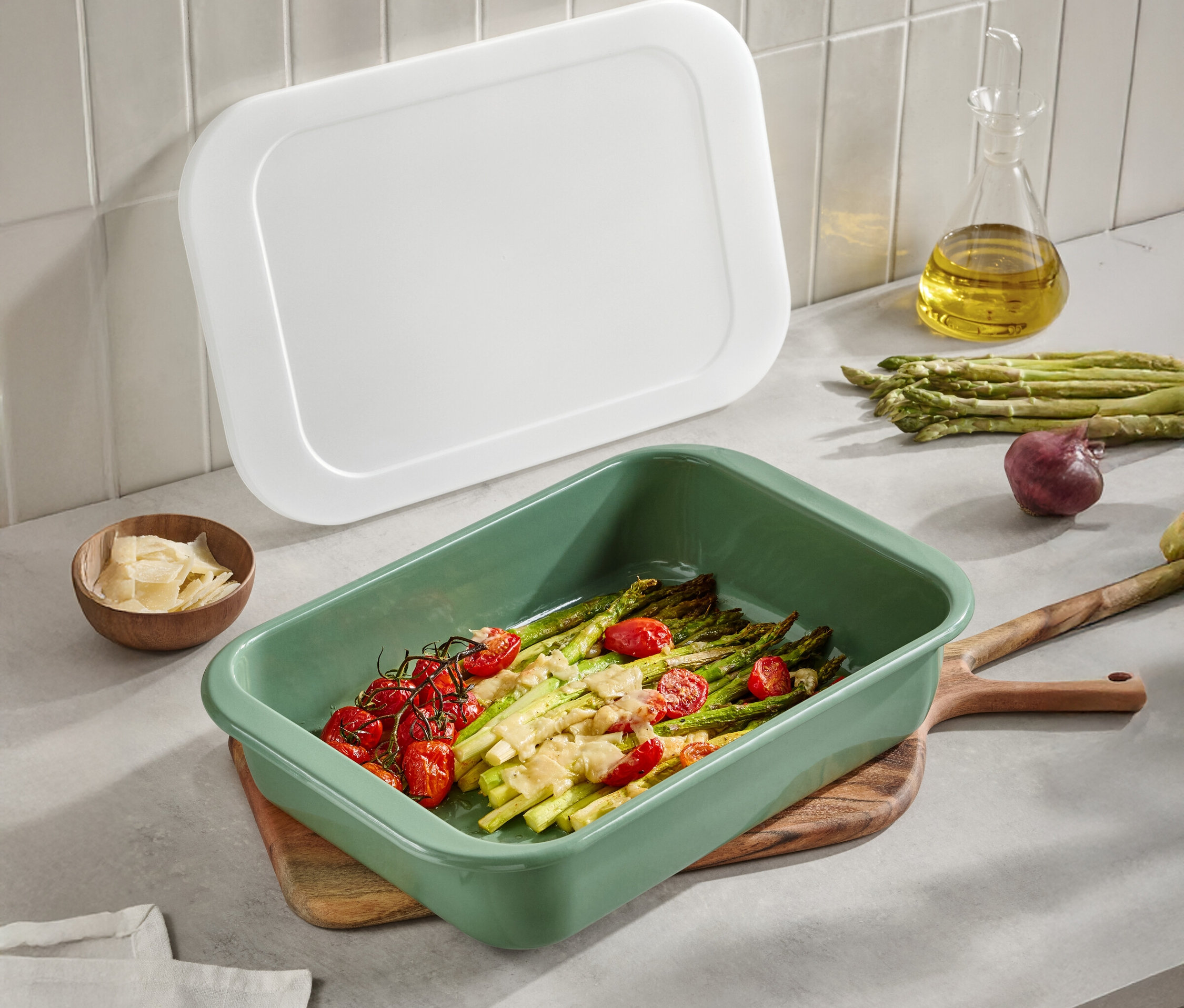 Zelená zapékací mísa s chřestem a rajčaty na dřevěném stole. Viditelné je také bílé víko, olivový olej, parmezán a cibule.