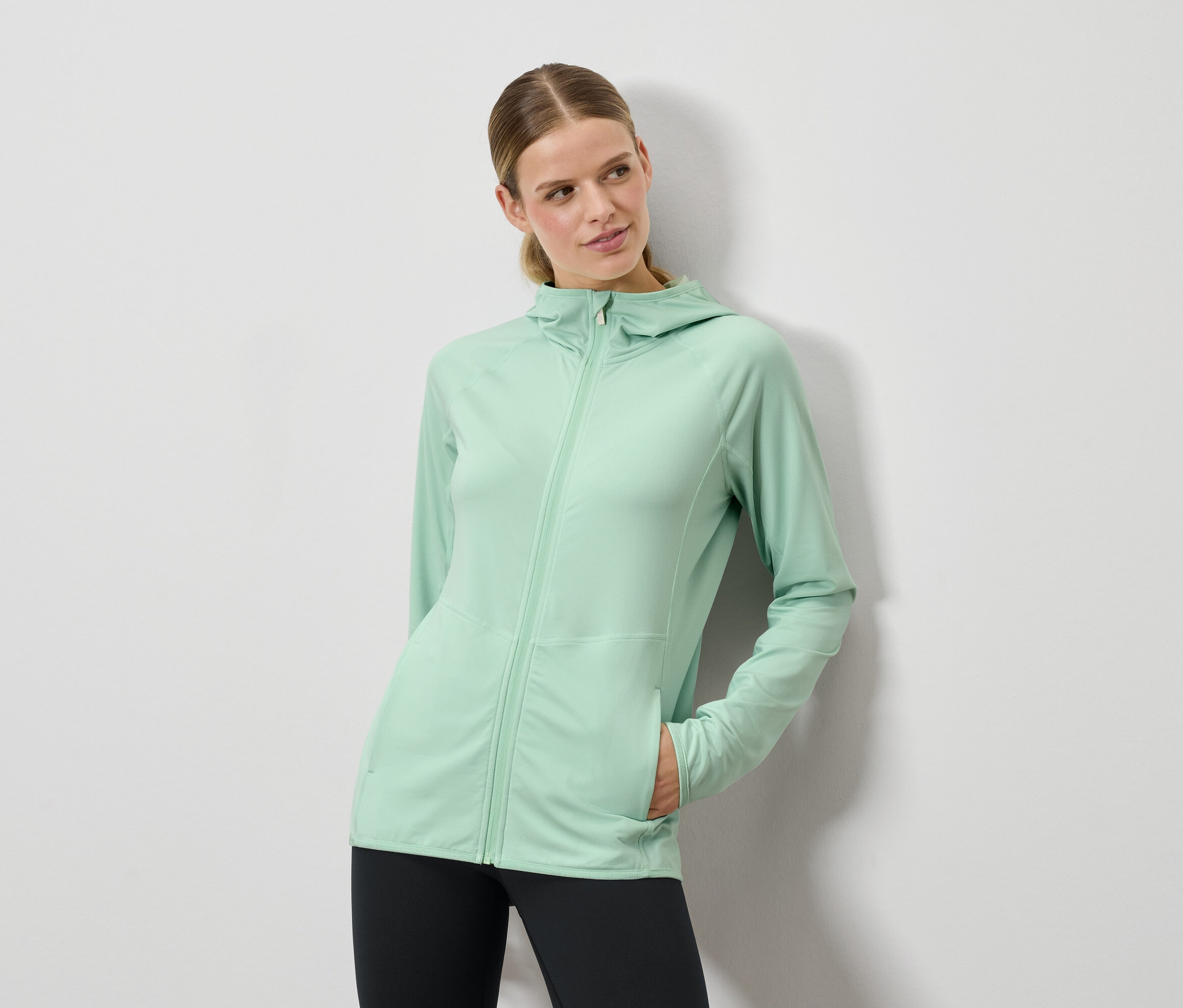 Žena pózuje v mátově zelném svetru s kapucí a černých legínách.