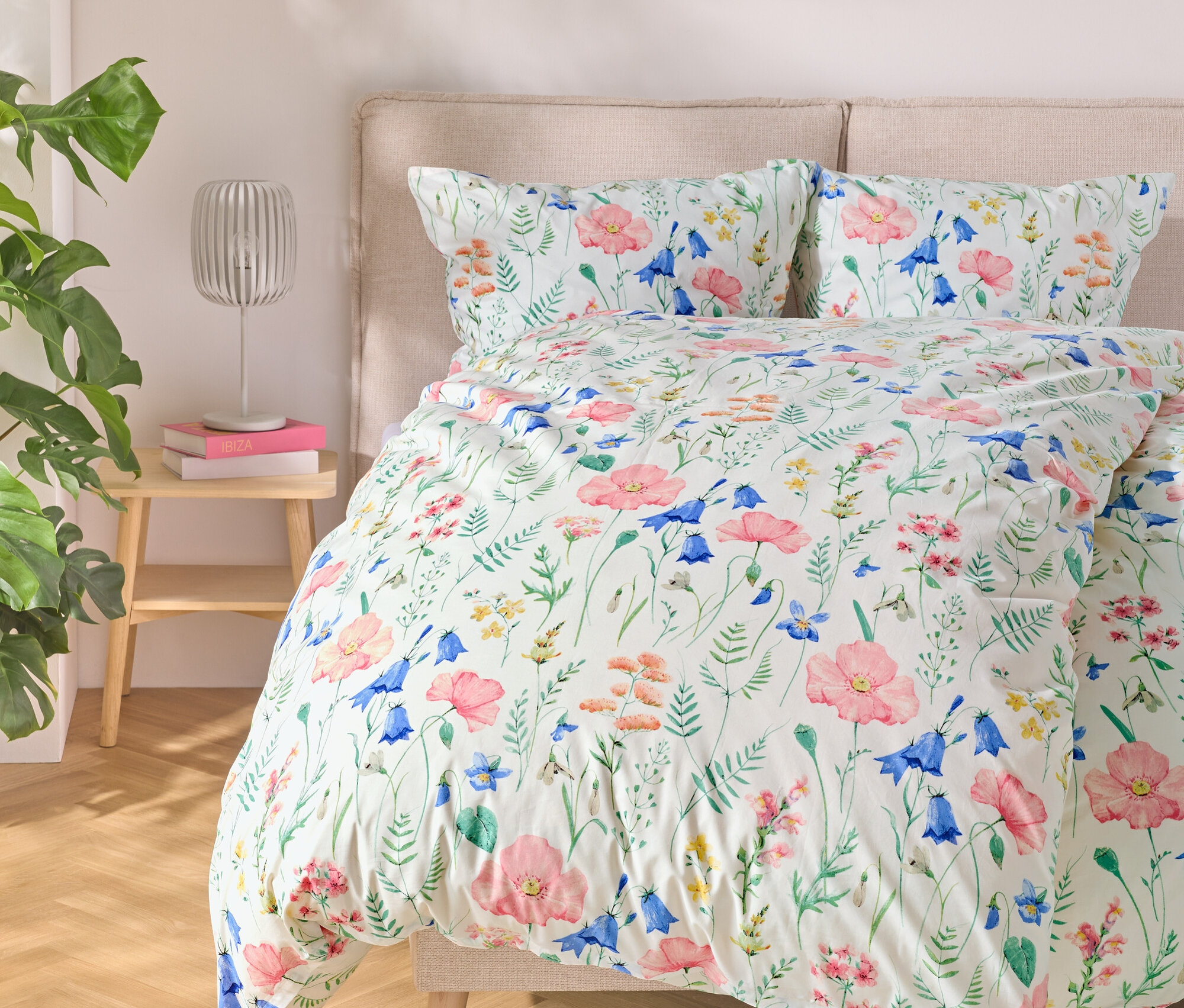 Postel s květinovým povlečením, u ní stolek s lampou a knihami a vlevo rostlina.