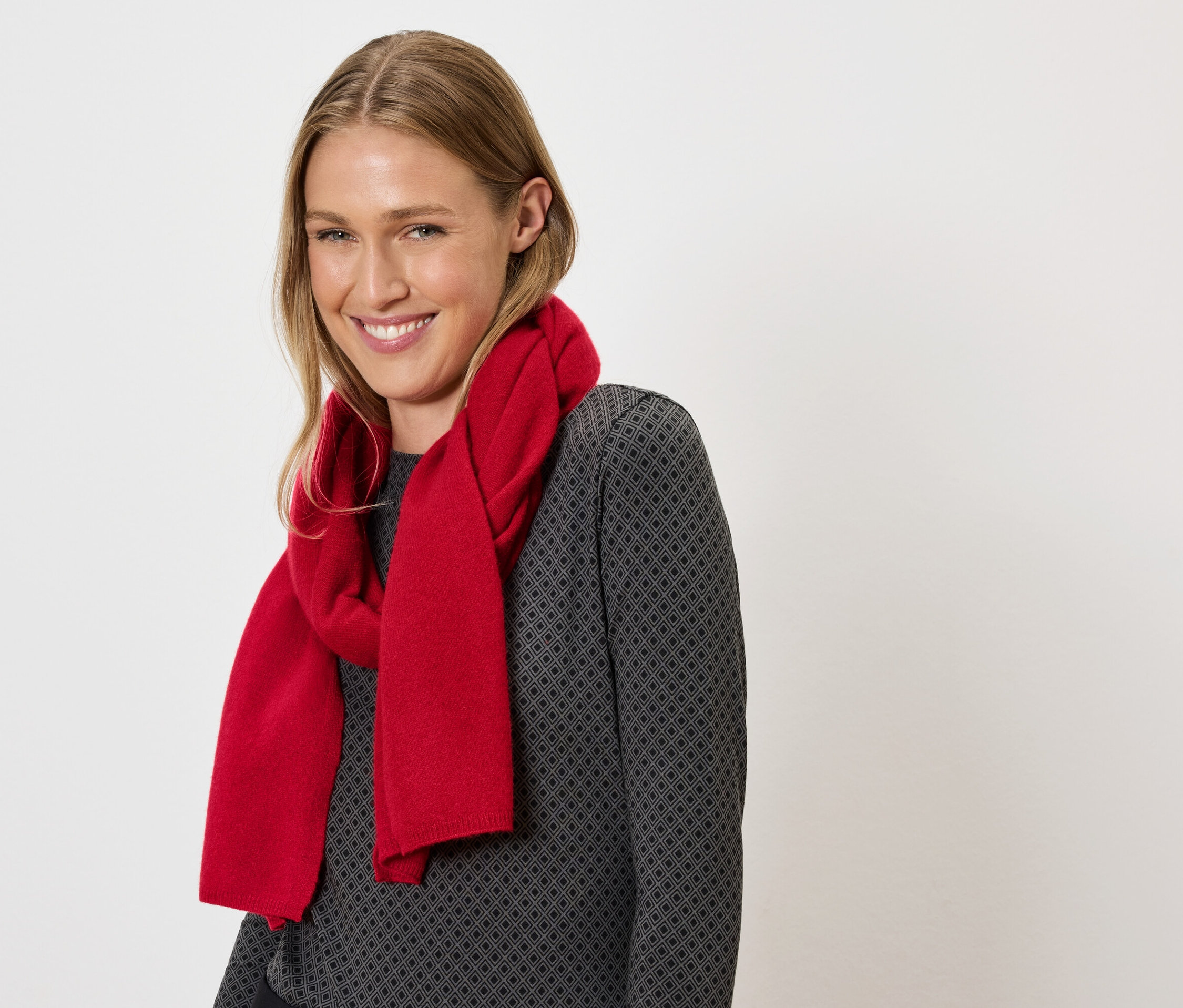 Usmívající se žena s blond vlasy a červenou šálou kolem krku na šedém svetru.