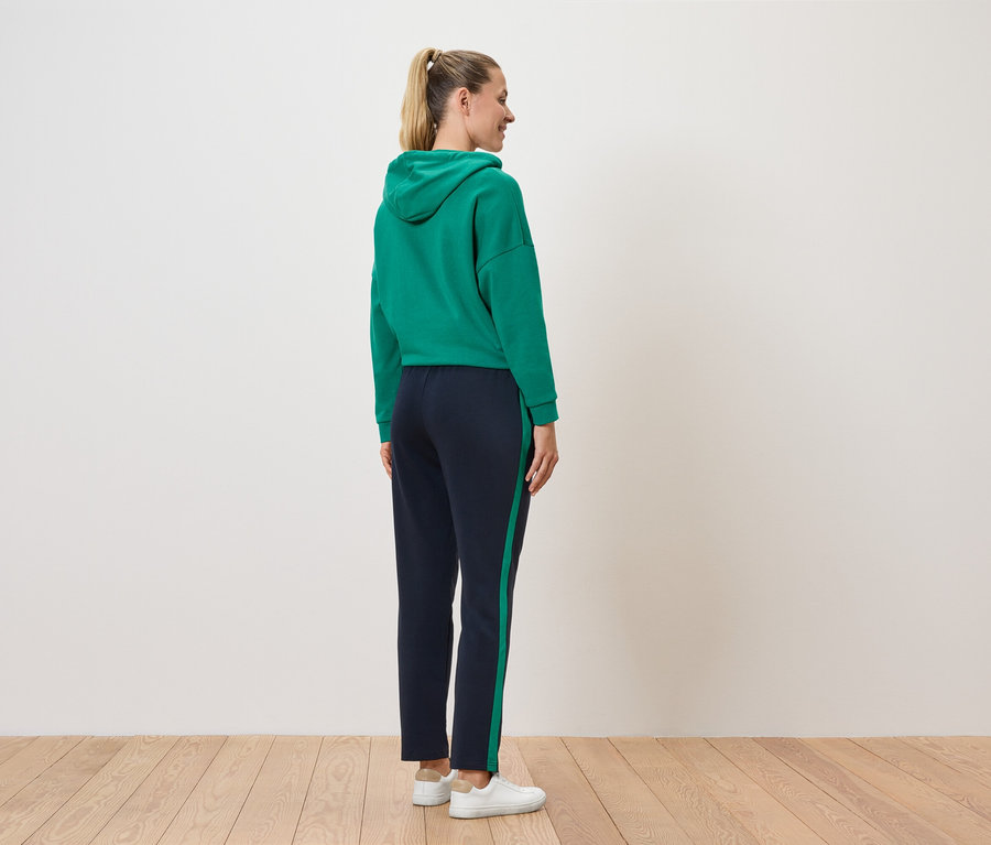 Žena stojí zády k divákovi a má na sobě zelenou mikinu s kapucí a tmavě modré kalhoty s proužkem.