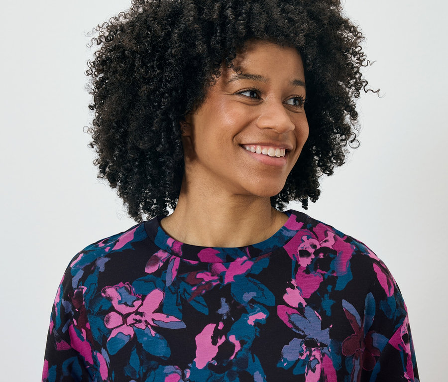 Portrét usmívající se ženy s afro účesem, oblečené v černém topu s květinovým vzorem v růžové a modré barvě.