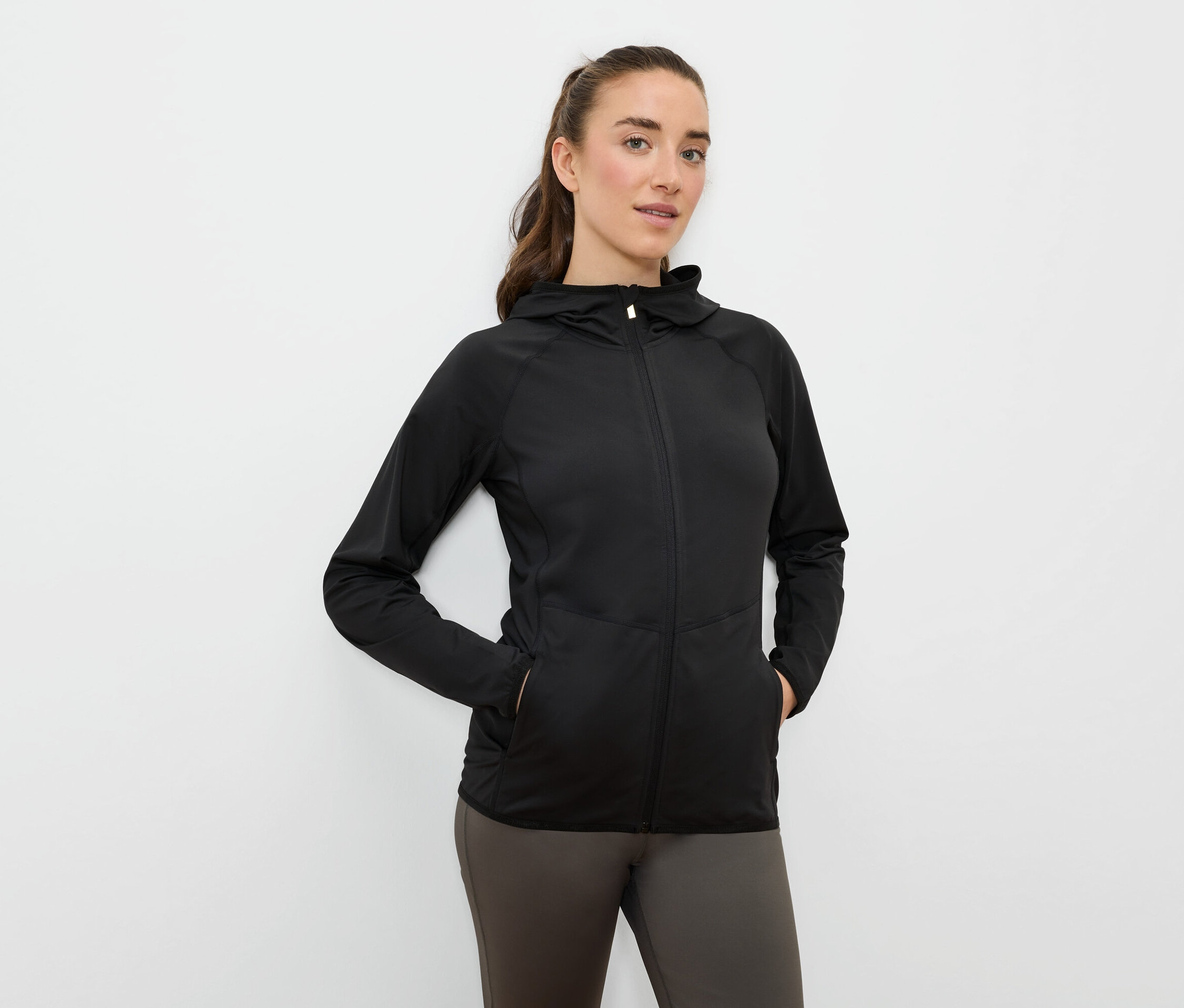 Žena má ruce v kapsách černé sportovní bundy s kapucí na zip.