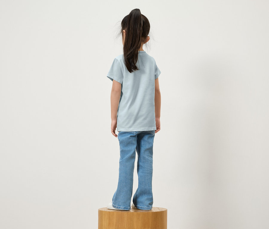 Zezadu dívka s culíkem stojí na dřevěném podstavci a má na sobě modré tričko a džíny.