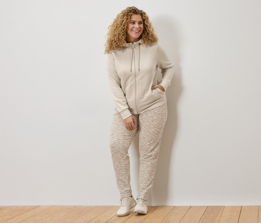 Žena s kudrnatými vlasy v béžové mikině s kapucí a kalhotách se zvířecím potiskem se opírá o bílou zeď.