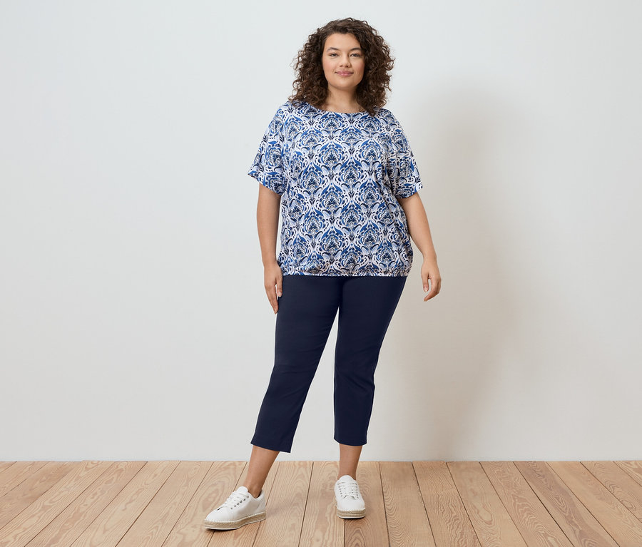 Žena s kudrnatými vlasy stojí před zdí a má na sobě modrou vzorovanou halenku, tmavě modré kalhoty a bílé tenisky.