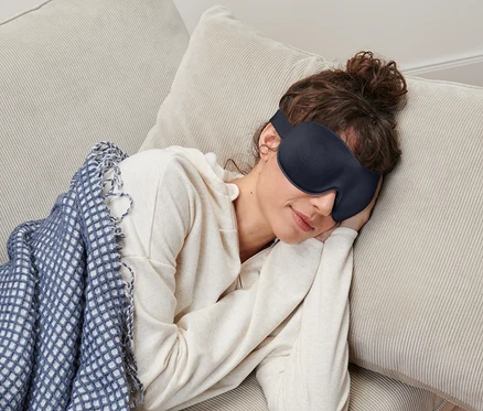 Žena s hnědými vlasy v drdolu s maskou na spaní leží na béžové pohovce pod modrobílou dekou.