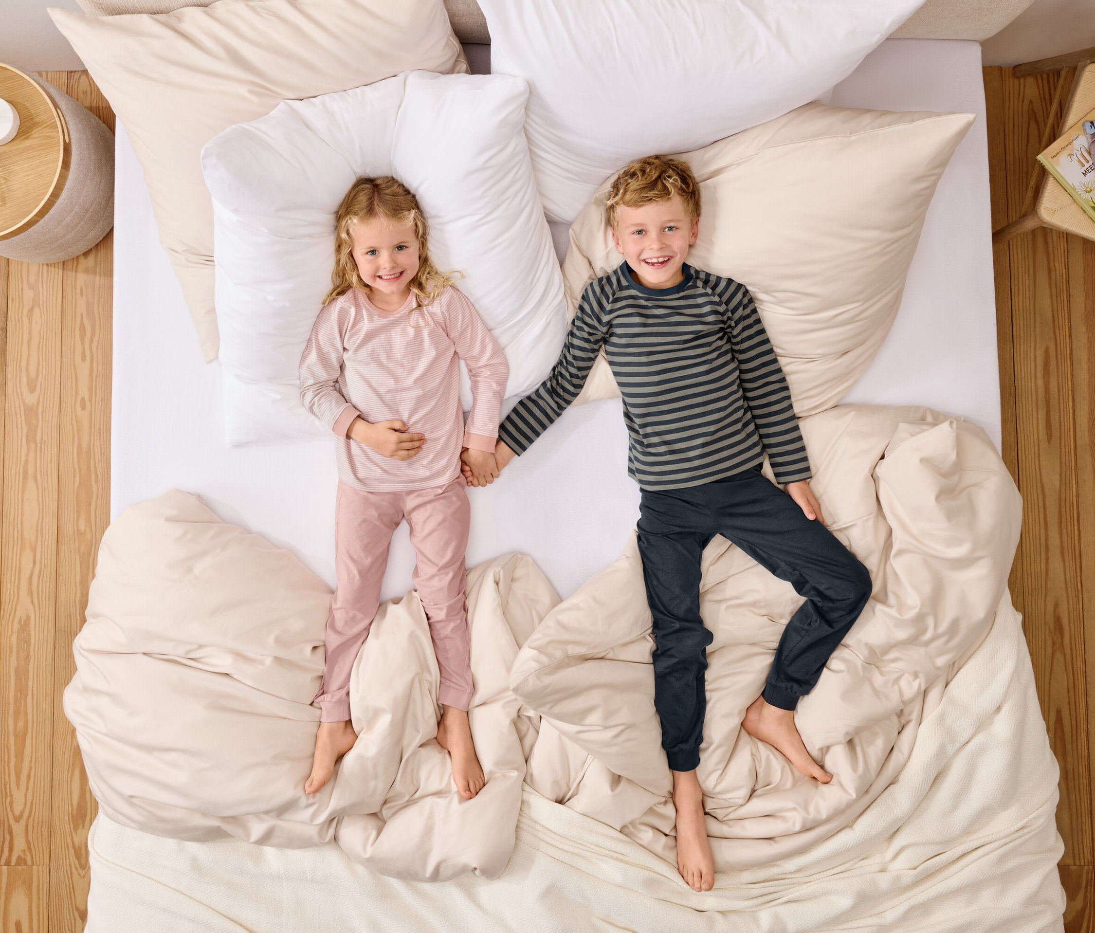 Pohled shora na dívku v růžovém pyžamu a chlapce v pruhovaném topu, kteří se drží za ruce na posteli s bílými polštáři.