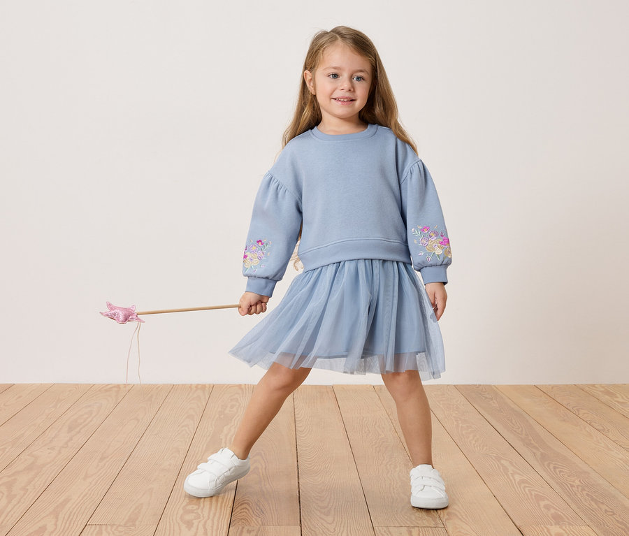 Dívka v modré mikině s květinovou výšivkou a tylové sukni, drží kouzelnou hůlku.