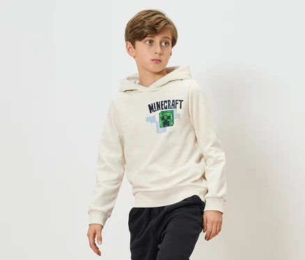 Chlapec má na sobě krémovou mikinu s kapucí Minecraft a černé tepláky na bílém pozadí.