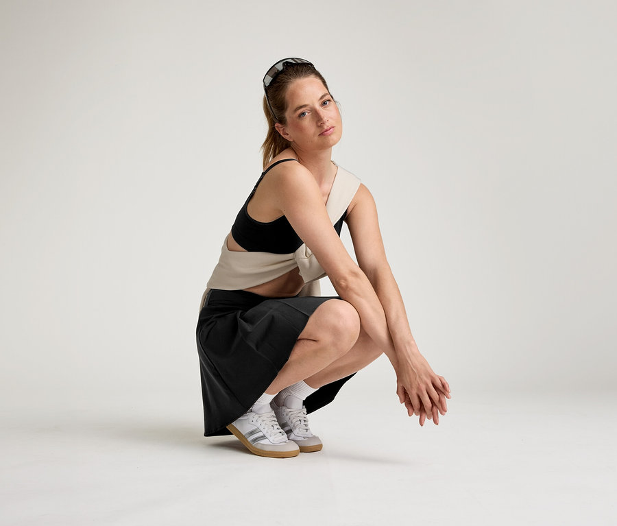 Žena se slunečními brýlemi na hlavě dřepí na bílém pozadí v černé sportovní podprsence, béžovém topu, černé sukni a bílých teniskách.