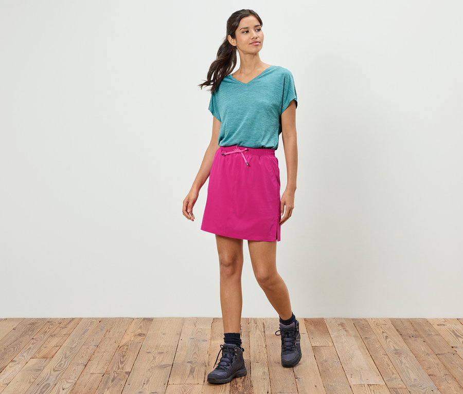 Žena kráčí v tyrkysovém tričku, růžové sukni a trekových botách po dřevěné podlaze před bílou stěnou.