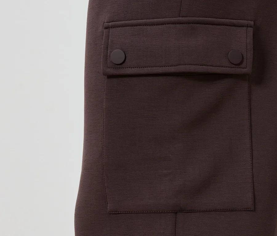 Detailní pohled na kaštanové kalhoty s našitou kapsou, která se zapíná na dva kulaté knoflíky.