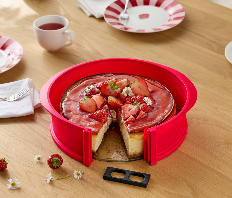 Jahodový cheesecake s červeným dortovým kruhem, nakrájený, na dřevěném stole s kopretinami, prostíráním a nápoji.