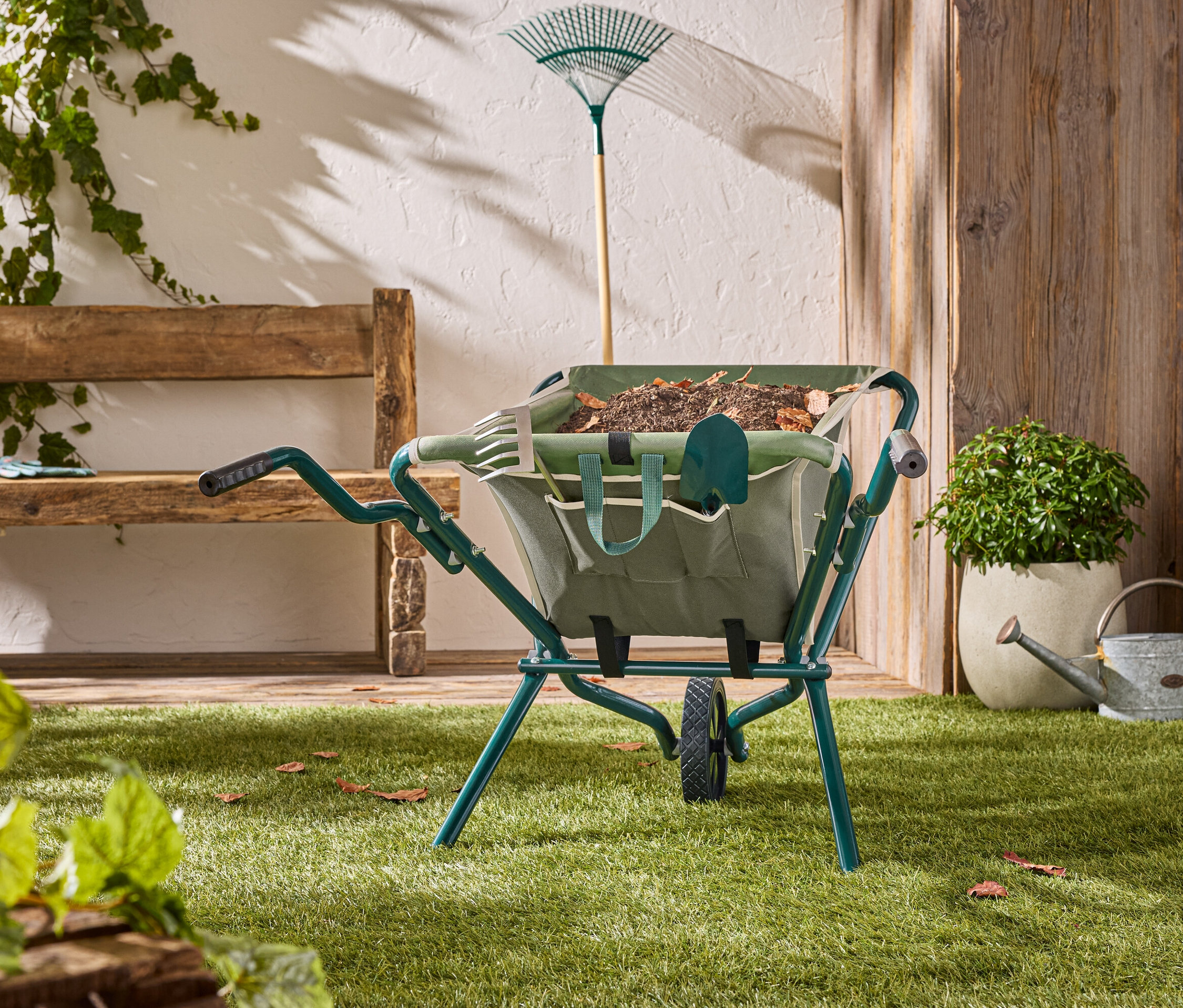 Zelený kolečko stojí na trávníku se zahradním nářadím uvnitř.