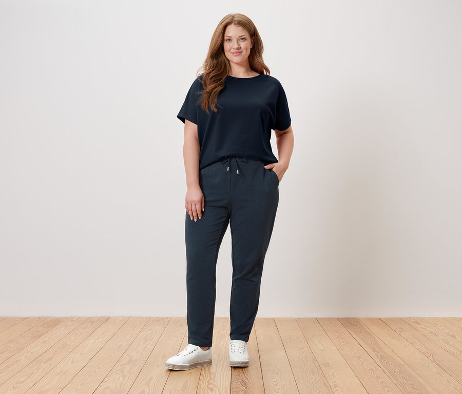 Žena stojí před zdí a má na sobě tmavě modré tričko, tmavě modré kalhoty a bílé boty.