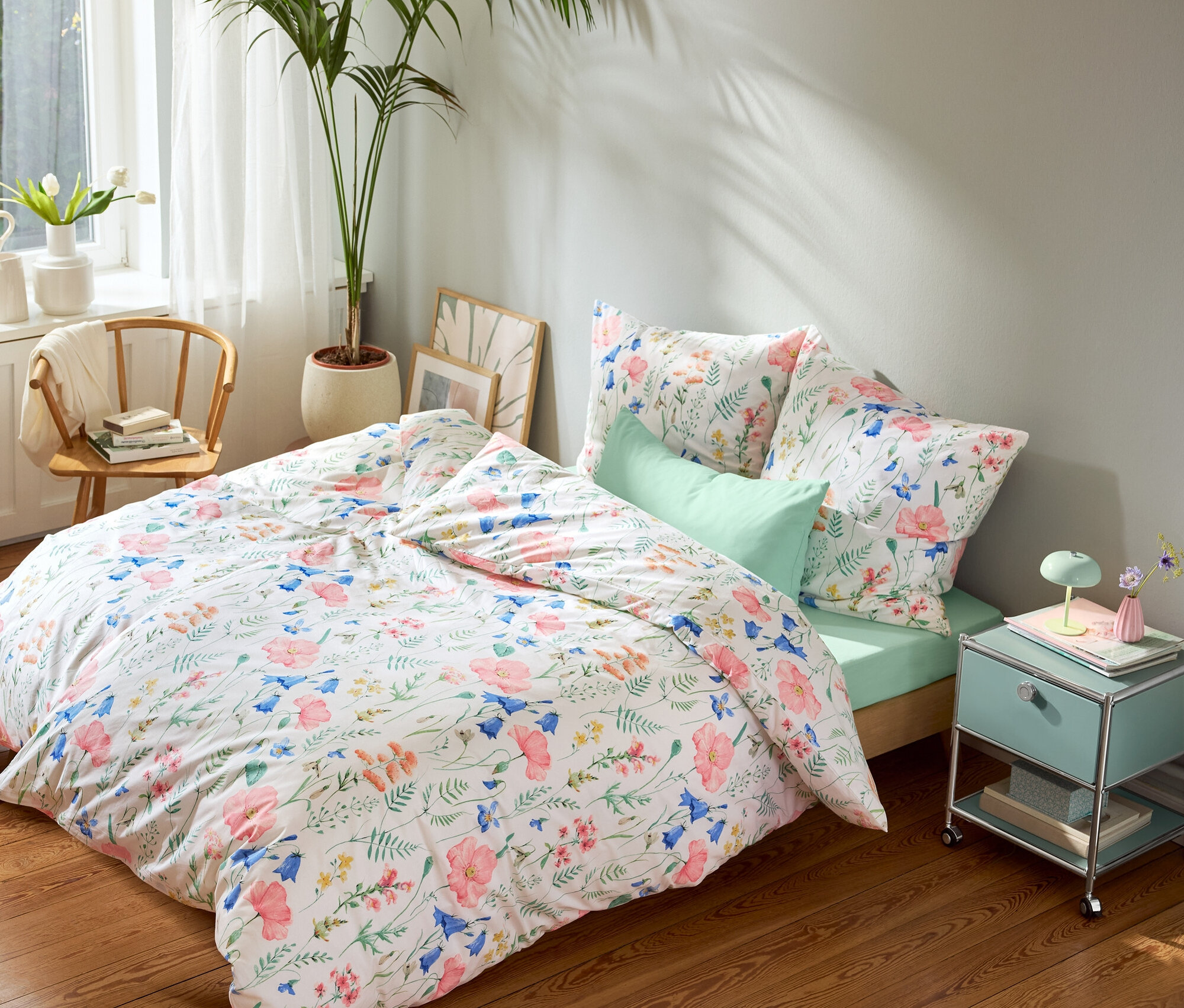 Postel s květinovým ložním prádlem, mátově zeleným prostěradlem a mátově zelenými povlaky na polštáře, mátově zelený noční stolek a kulatý koberec.