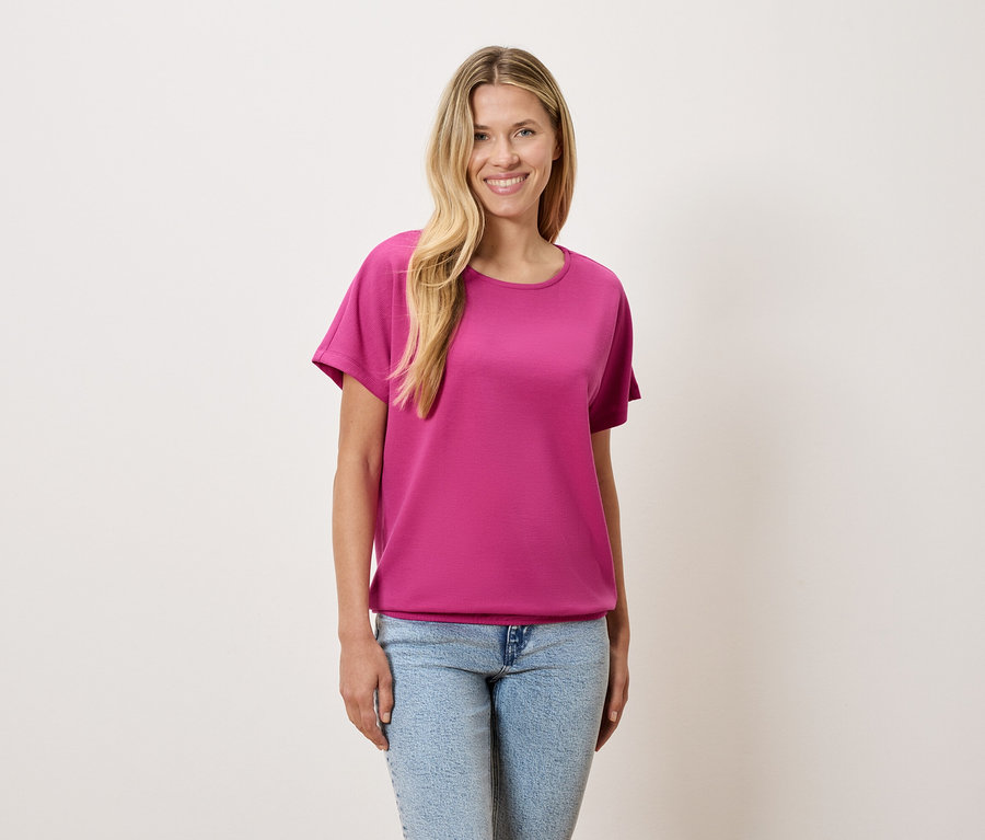 Žena s blond vlasy má na sobě růžové tričko a modré džíny.
