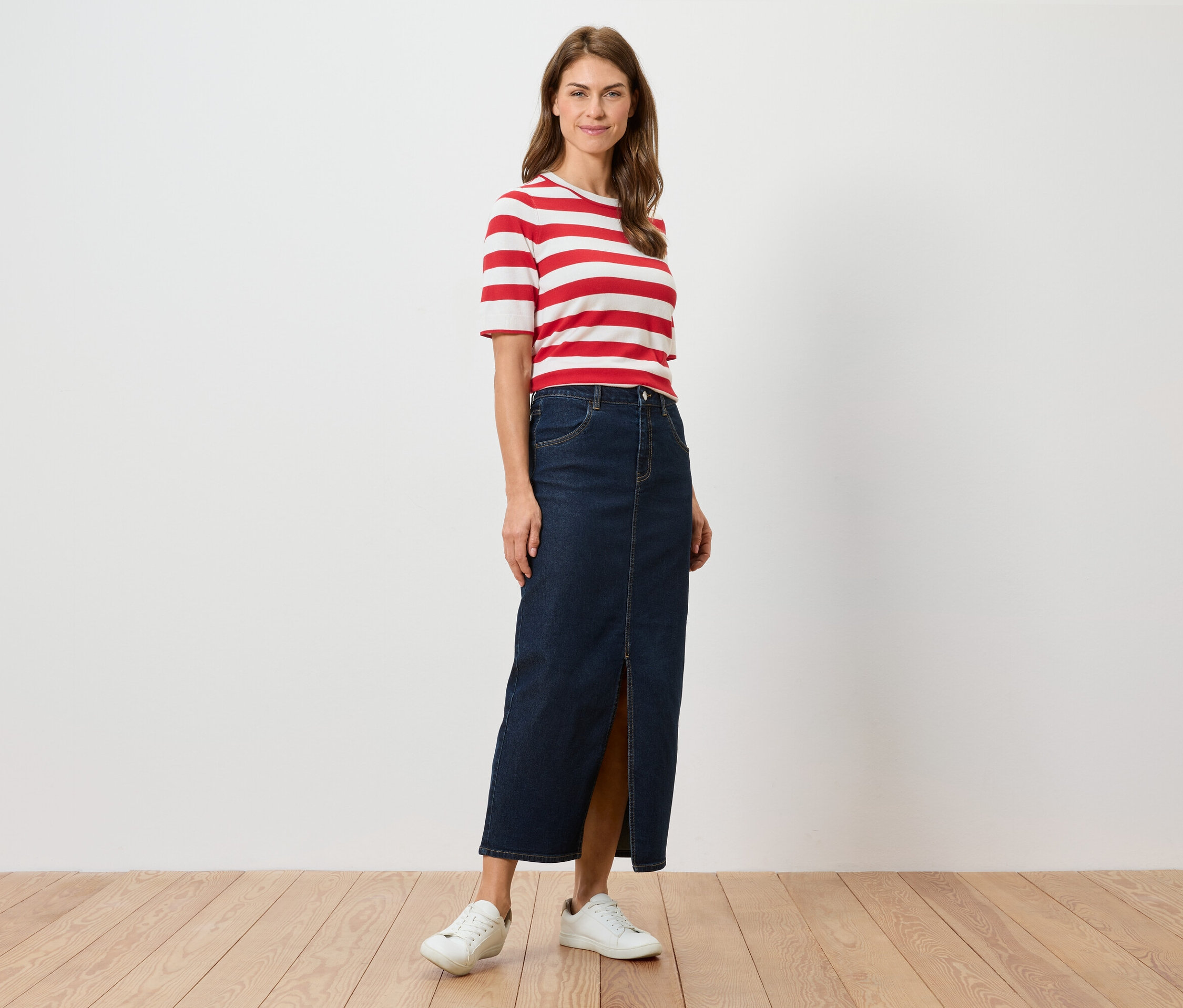 Žena stojí a má na sobě červeno-bílé pruhované tričko, dlouhou džínovou sukni s rozparkem a bílé tenisky.