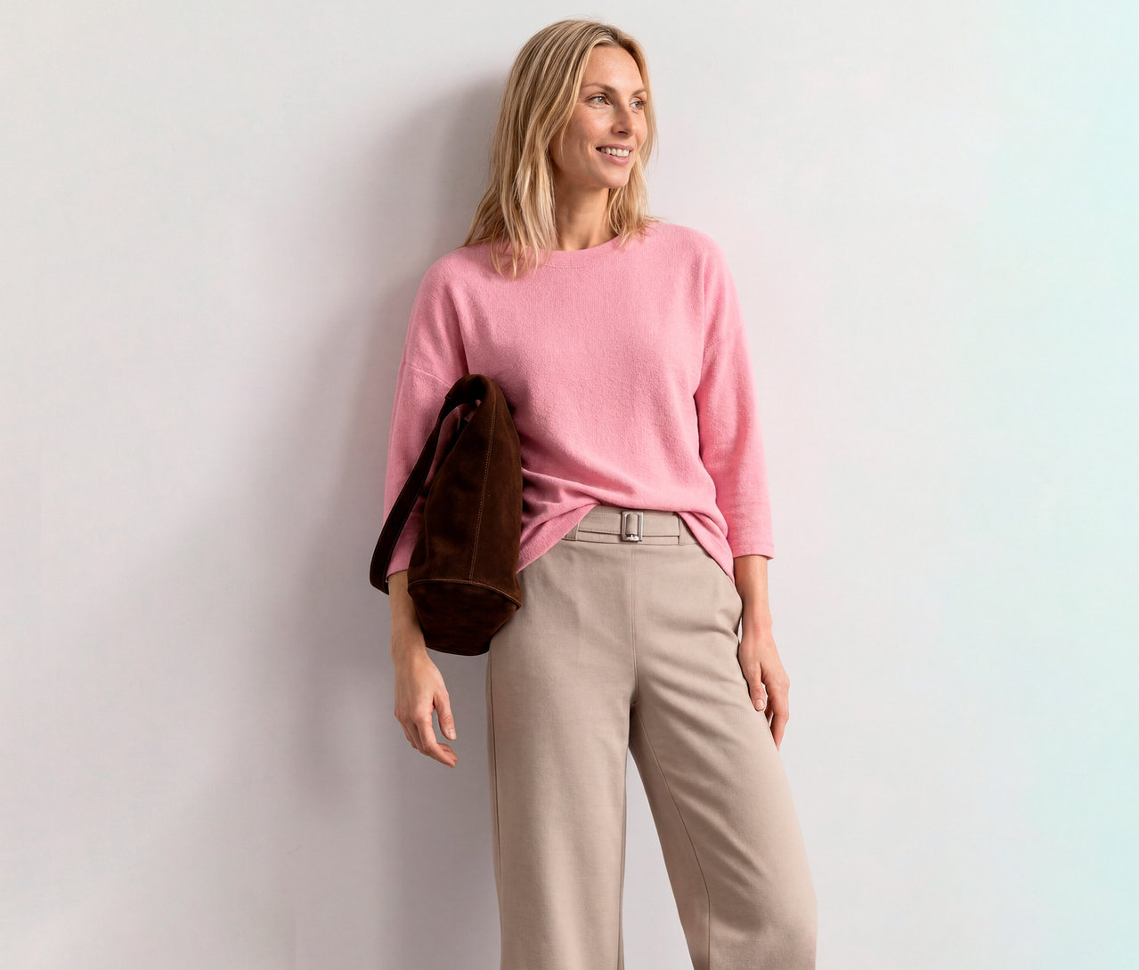 Usmívající se blondýnka v růžovém triku s 3/4 rukávem Street One, béžových kalhotách a hnědé tašce, opřená o bílou zeď.