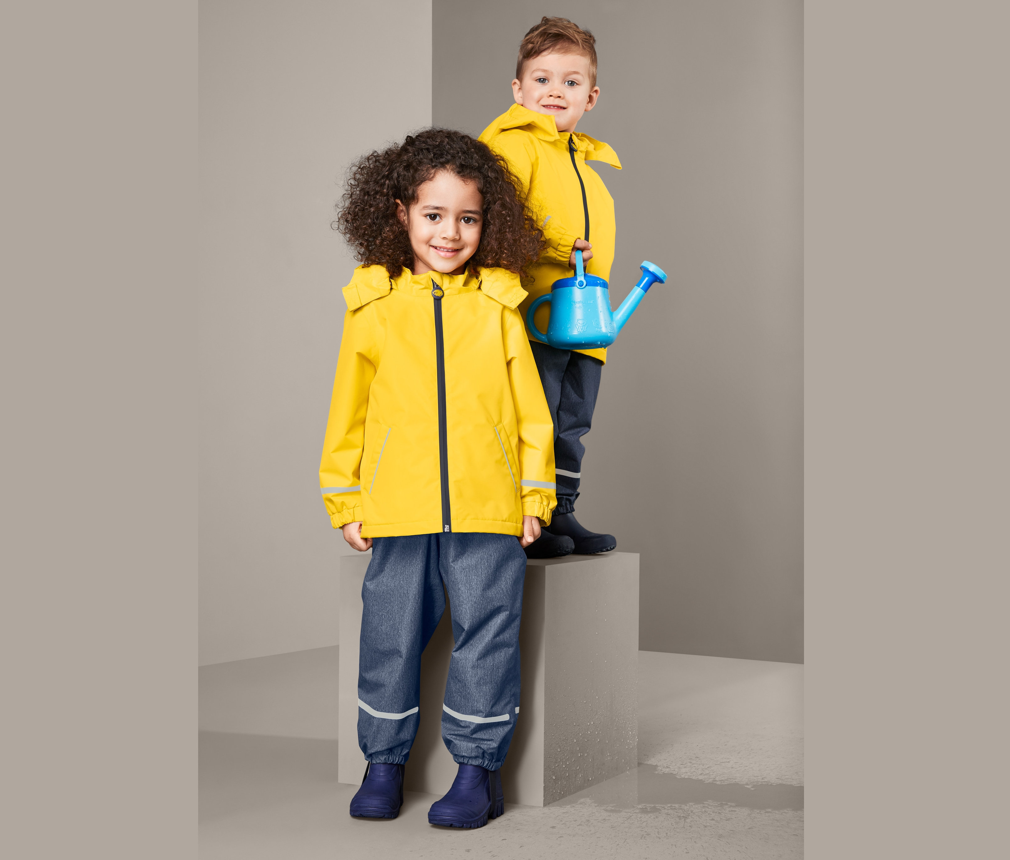 Dvě děti pózují: dívka v termobundě do deště z recyklovaného materiálu a chlapec držící modrou konev.
