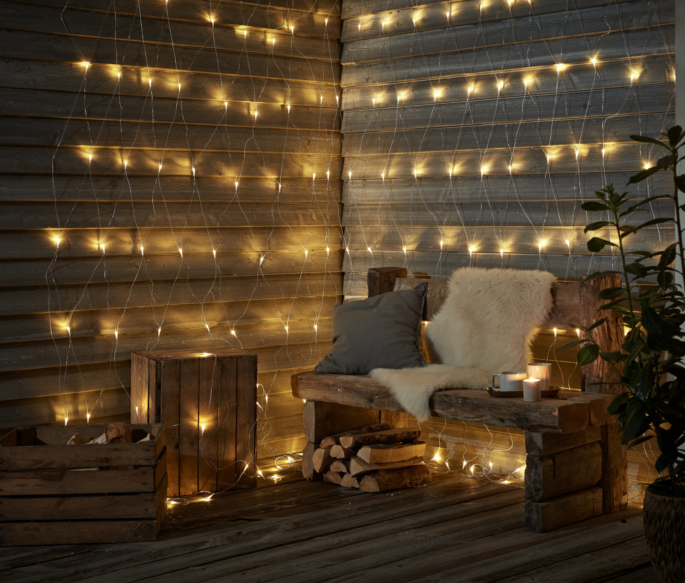 Dřevěná lavička s polštáři a kožešinou stojí před stěnou zdobenou venkovní světelnou sítí s LED.