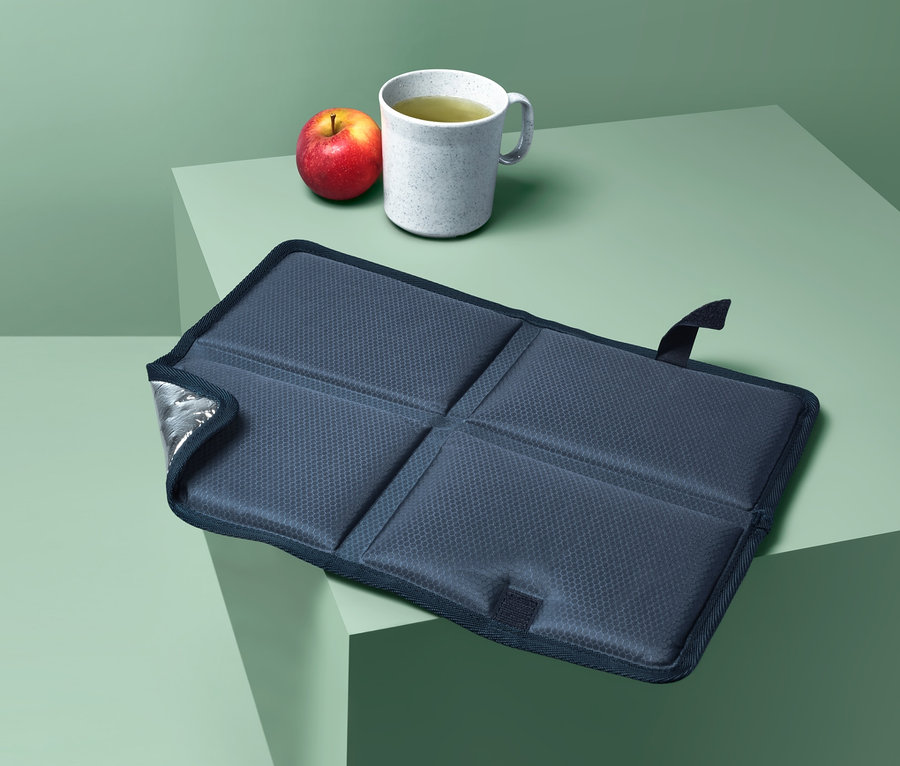 Skládací termopolštářek na sezení vedle jablka a hrnku na zeleném stole.