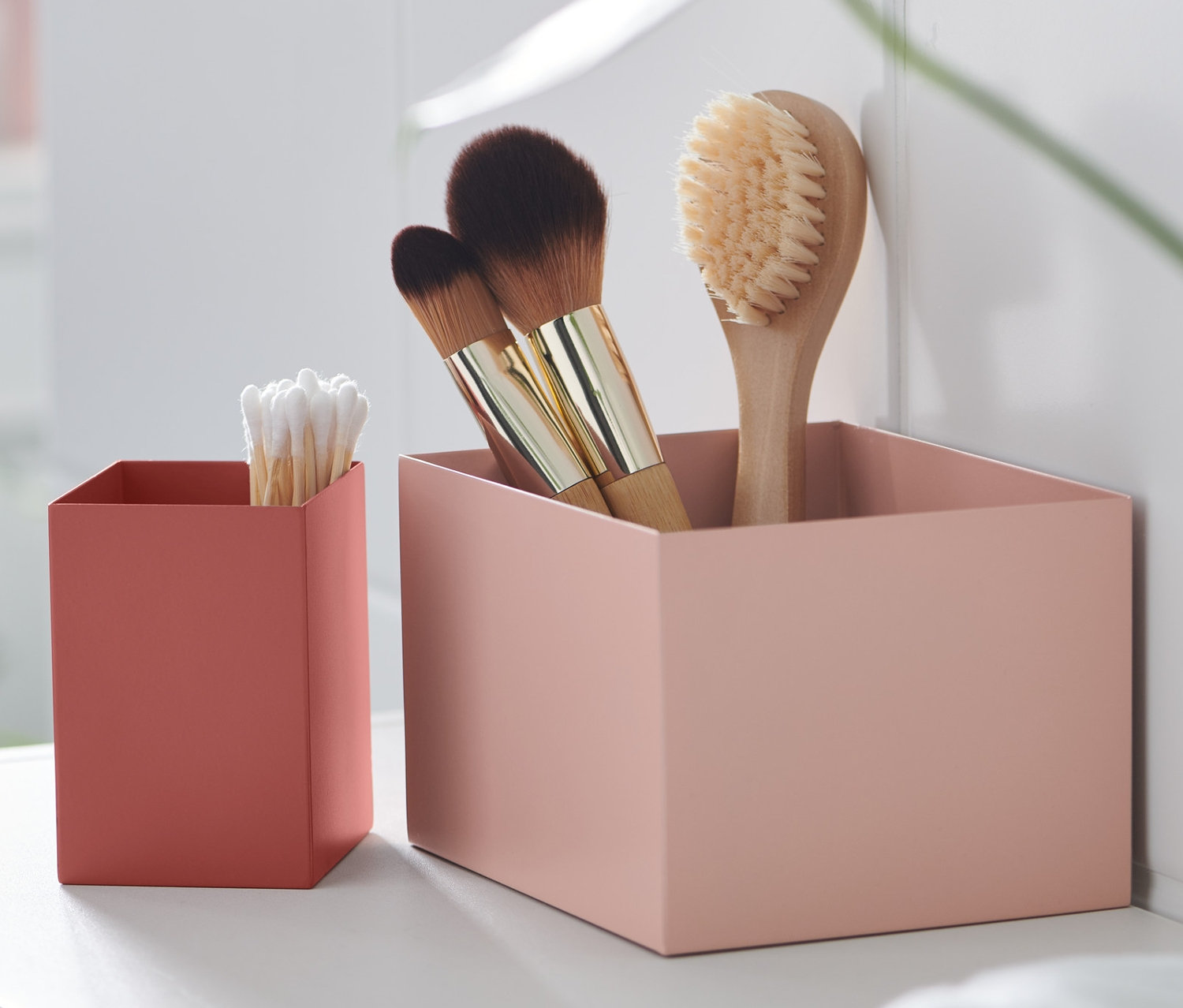 Dvě dekorativní kovové krabice na bílé polici, v jedné štětce a v druhé vatové tyčinky.