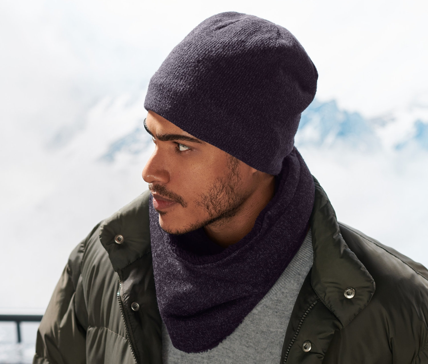 Muž s pletenou modrou kruhovou šálou z fleecu a čepicí na sobě, v pozadí zasněžené hory.