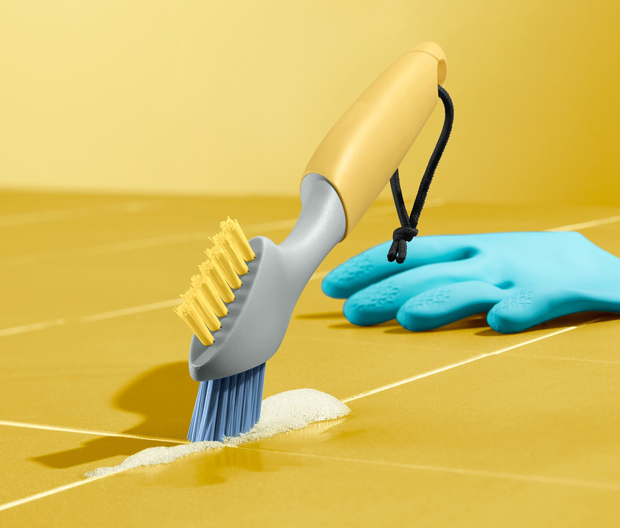 Kartáč na spáry čistí žluté dlaždice. Vedle leží modrá rukavice.