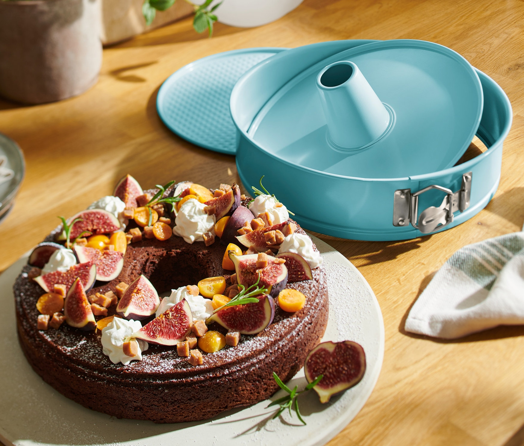 Čokoládová bábovka s fíky na talíři vedle modré formy na dort s přepravním víkem.