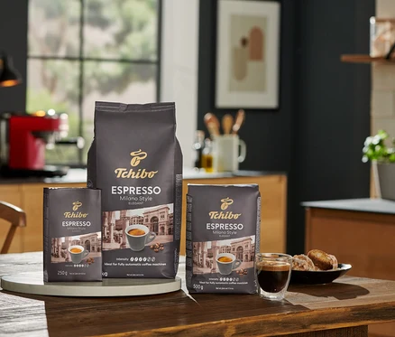 Tři balení zrnkové kávy Tchibo Espresso Milano Style 250 g, 500 g a 1 kg na dřevěném stole v kuchyni.