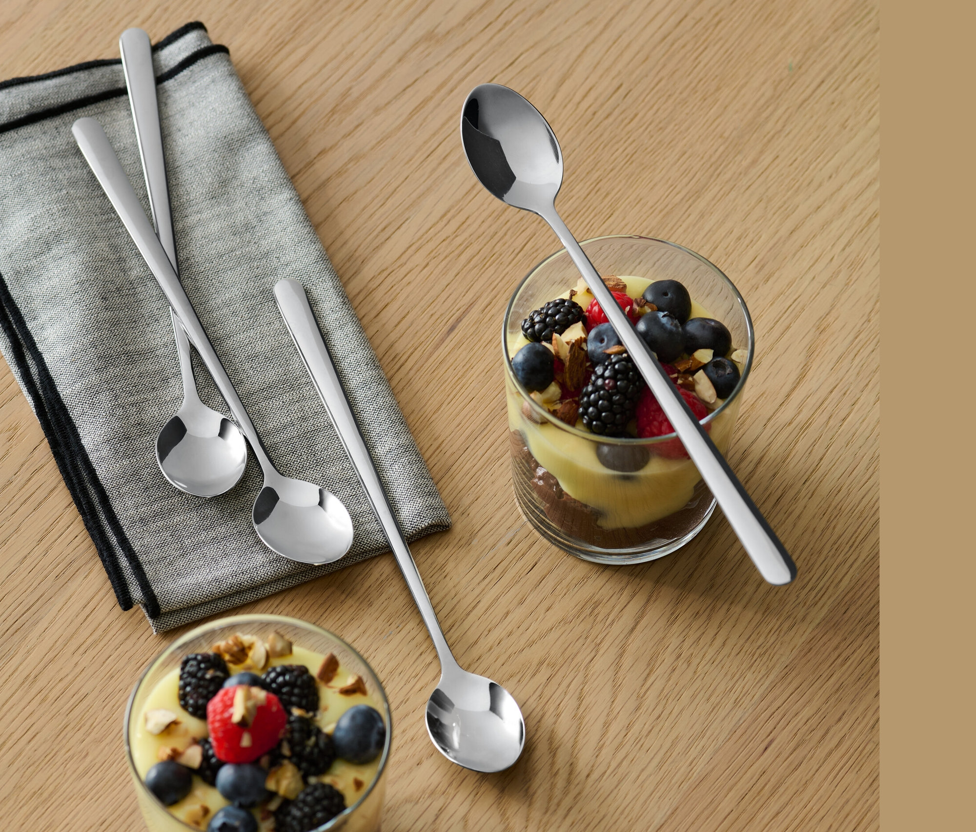 Na dřevěném stole leží šedá utěrka a dvě sklenice s dezerty a ovocem. U každé sklenice leží jedna ze 4 dlouhých lžiček.