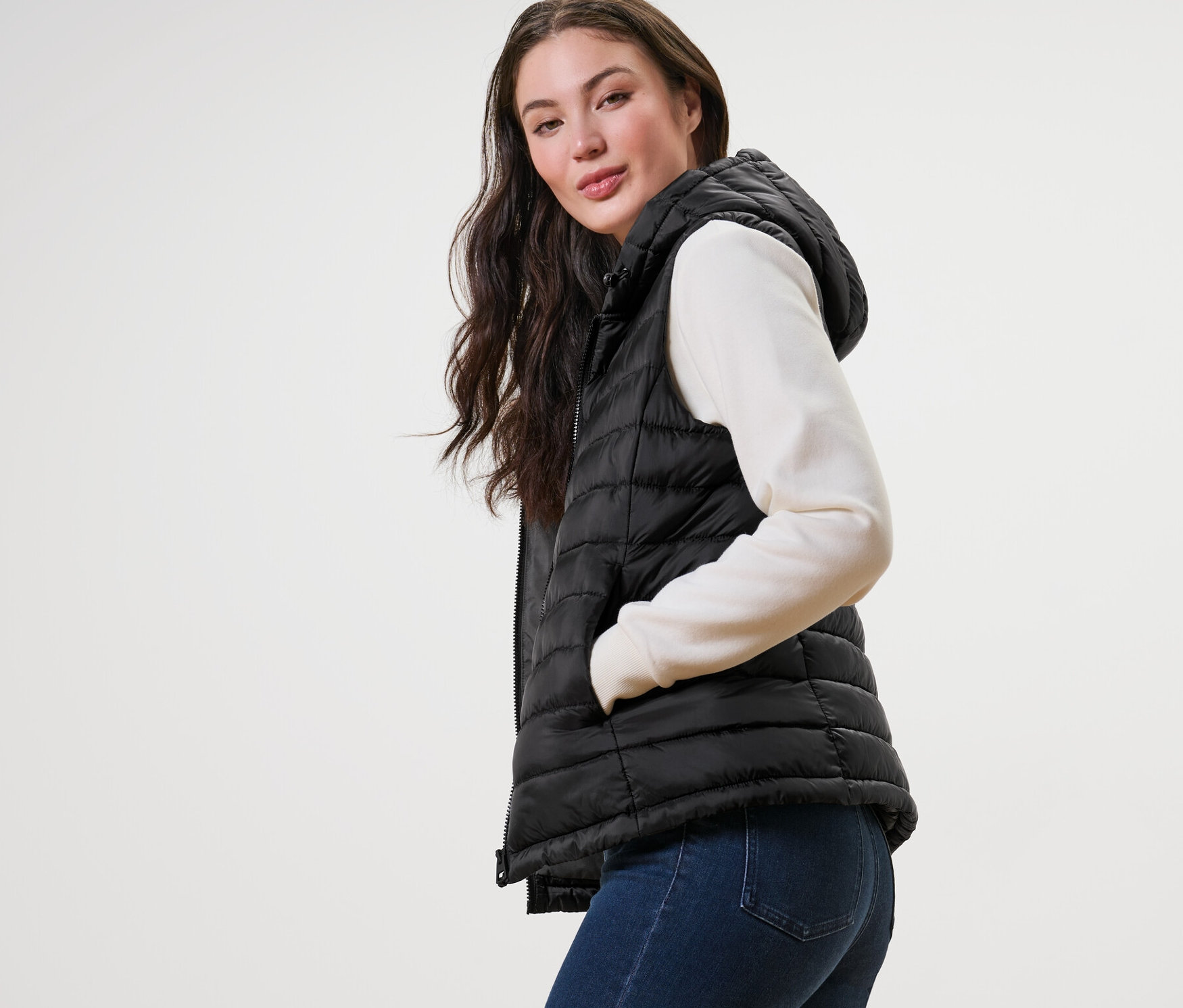 Žena pózuje v prošívané vestě s kapucí, svetru a džínách.