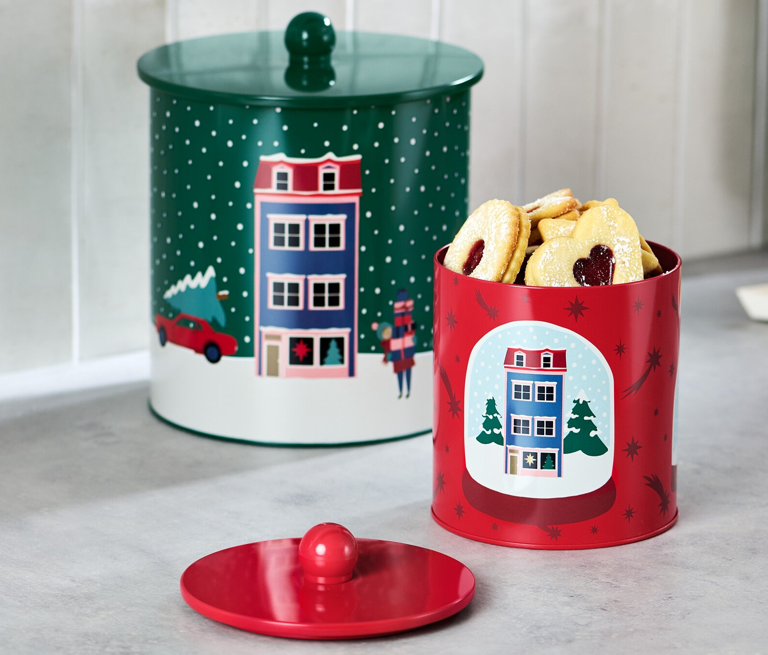 Dvě dózy na cukroví stojí na lince. Jedna dóza je zelená s víkem a druhá červená s sušenkami.