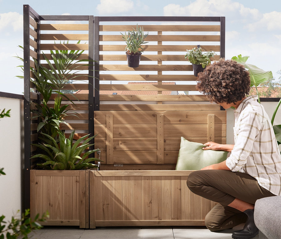 Žena pokládá polštář na boční díl zástěny »Elin« s lavicí s úložným prostorem. V pozadí je rohový díl zástěny »Elin« s truhlíkem.