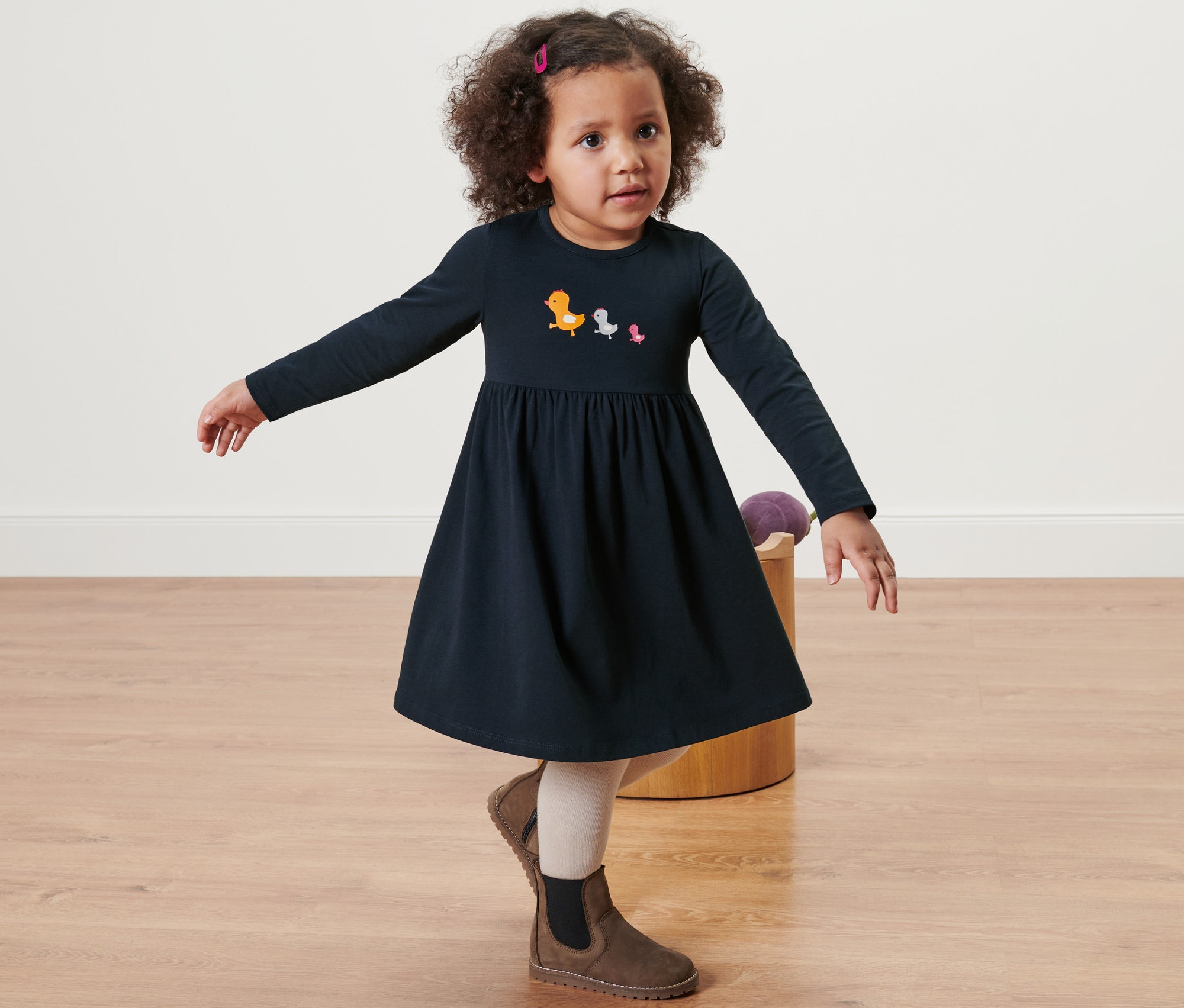 Holčička pózuje v modrých žerzejových šatech s dlouhým rukávem a obrázky ptáčků, doplněných hnědými botami.