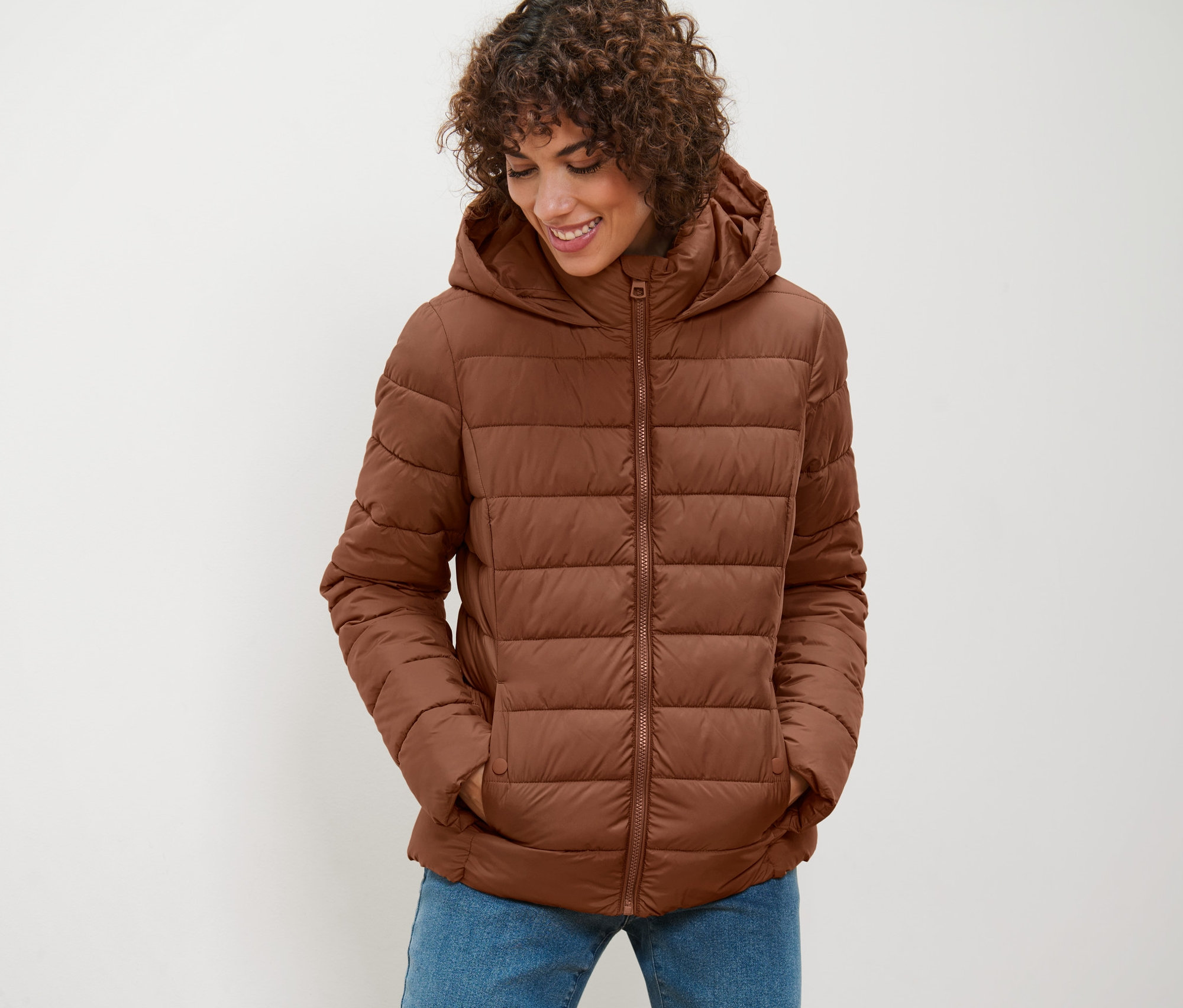 Žena má na sobě koňakovou prošívanou bundu s kapucí a dívá se dolů.