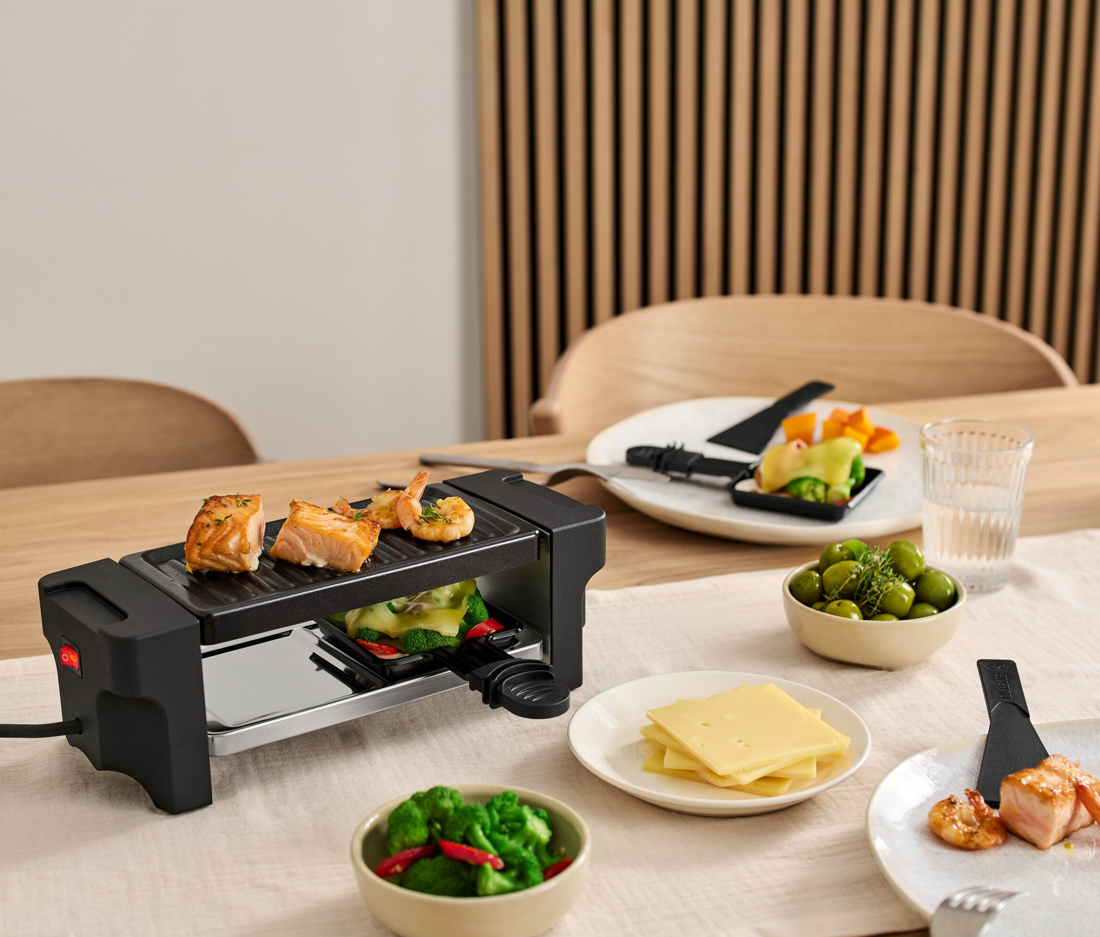 Na stole je raclette gril pro 2 osoby s lososem a krevetami na grilu. Talíře s ingrediencemi jsou kolem grilu.