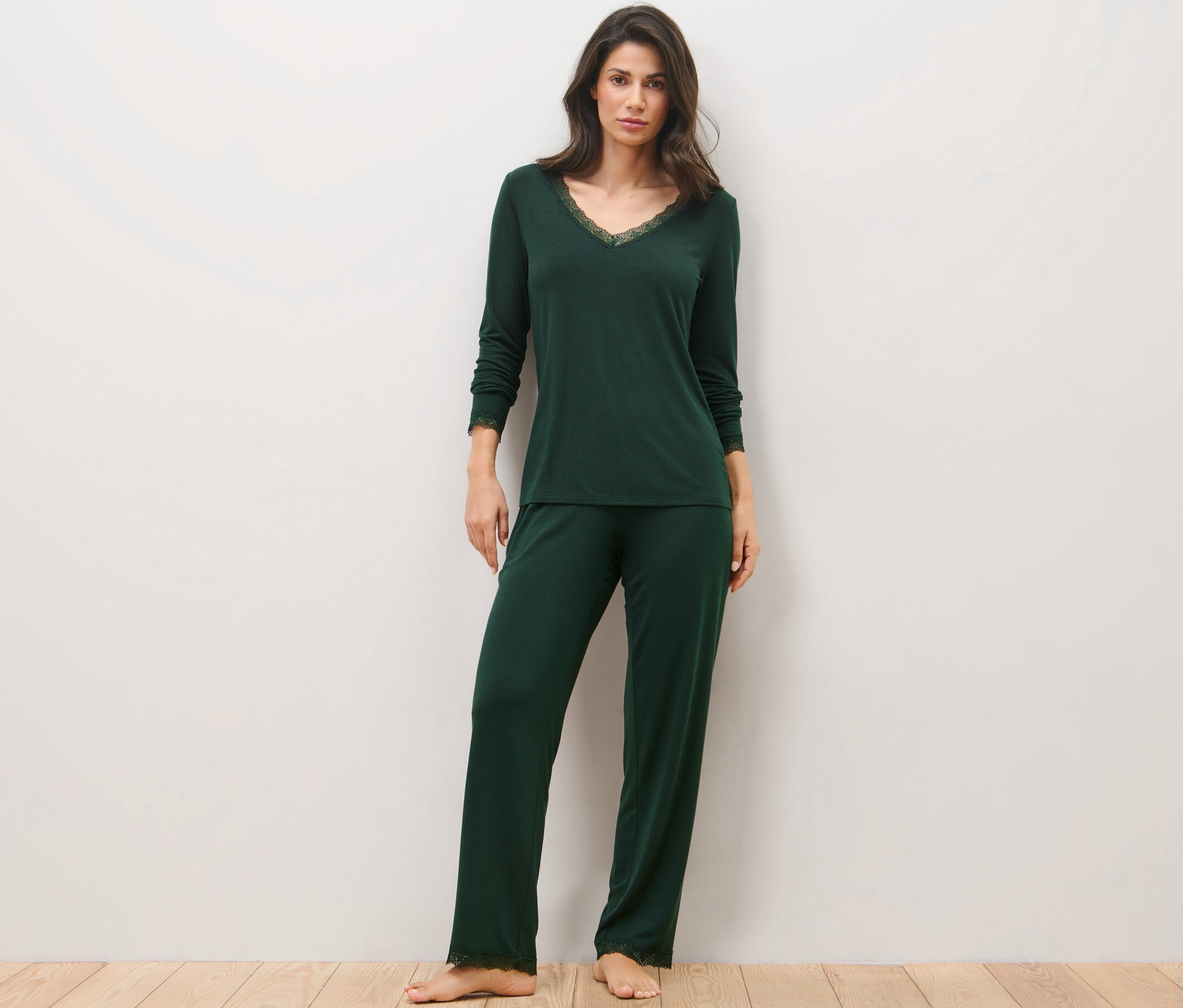 Žena stojící na dřevěné podlaze má na sobě tmavě zelené pyžamo s krajkovým lemováním.