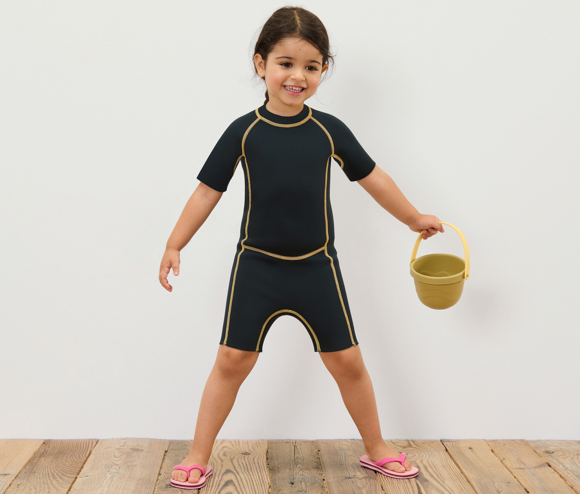 Dívka v dětském neoprenovém obleku drží žlutý kbelík.