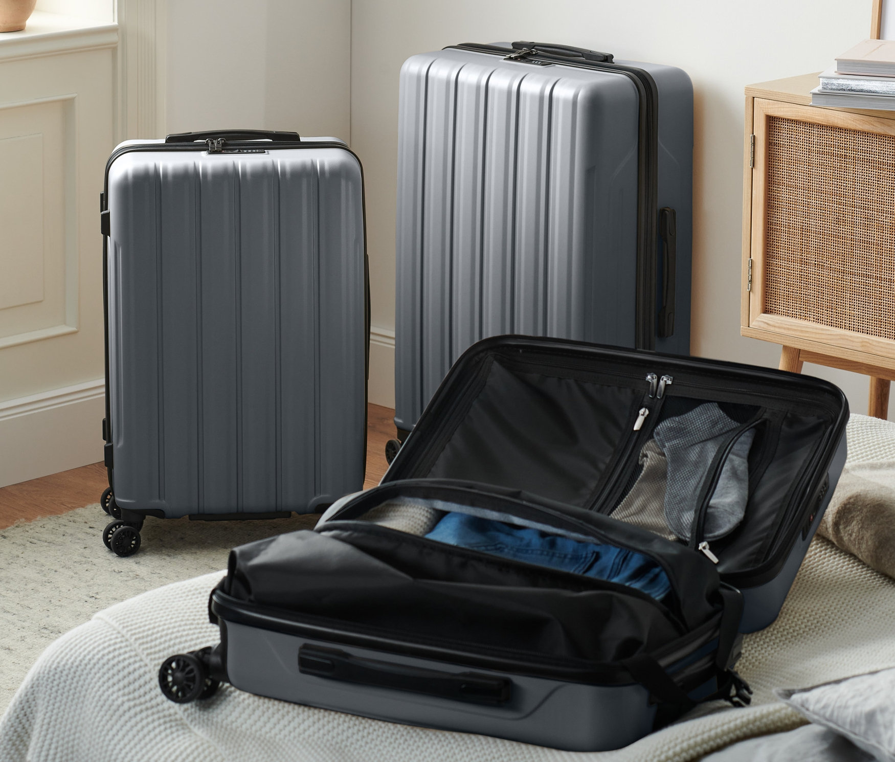 Tři šedé kufry z tvrdé skořepiny (malý, střední a velký) stojí v interiéru, jeden je otevřený na posteli.