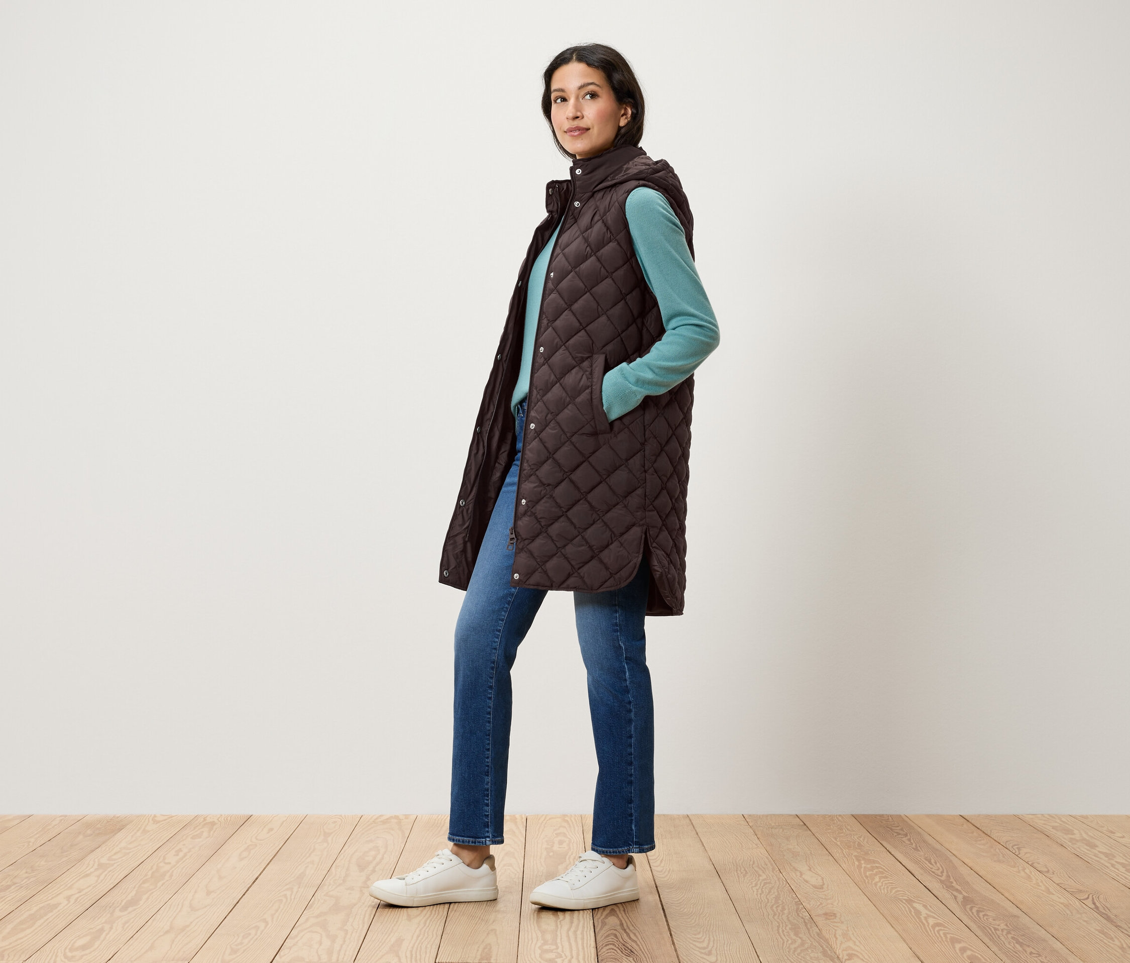Žena pózuje v prošívané hnědé vestě s kapucí, modrém svetru, džínách a bílých botách.