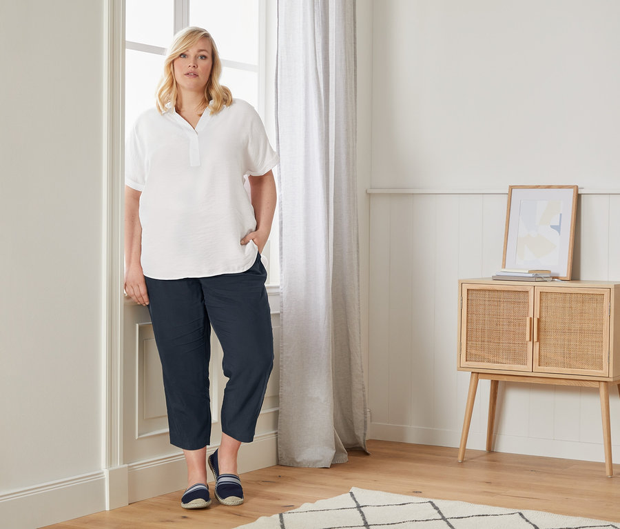 Žena pózuje u okna v halence ve stylu oversize, ležérních kalhotách chino a espadrilkách.